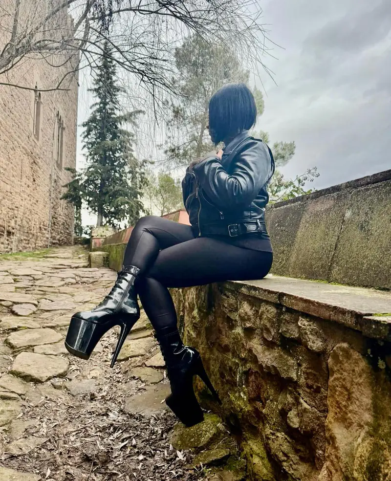 Miss Soniax sentada en un muro de piedra junto a un castillo, con chaqueta de cuero y botas de plataforma